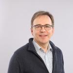 Harmaaseen vetoketjulliseen neuleeseen pukeutunut Arto katsoo hymyillen suoraan kameraan.