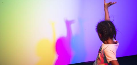 Child holding one hand up, in front of child is colorful wall.
