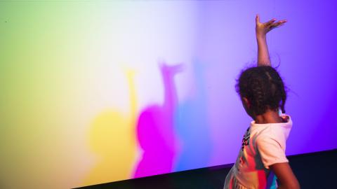 Child holding one hand up, in front of child is colorful wall.