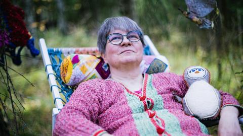 Henkilö istuu lepotuolissa puutarhassa. Hänellä on kainalossaan itse tehty käsityöpehmolelu.
