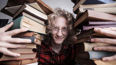 Kiharatukkainen nuori mies istuu ja nojaa samalla pöytään. Hän on kahmaissut syleilyynsä kaksi iso pinoa vanhoja kirjoja.  