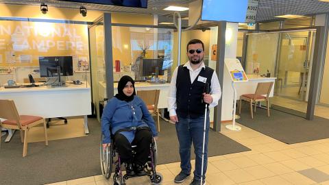 Liikuntavammainen nainen istuu pyörätuolissa. Hänen oikealla puolellaan seisoo näkövammainen mies, jolla on kädessä valkoinen sokean keppi. Molempien kasvot ovat kohti kameraa.