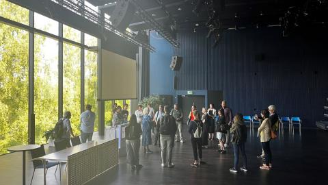 Ihmisiä kuljeskelemassa esitystilassa, jossa on suuret lasi-ikkunat koivuja päin. 
