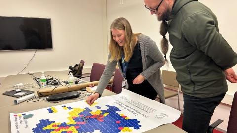 Nainen ja mies ovat kumartuneet pöydän ylle tarkastelemaan käsitekarttajulistetta kokoushuoneessa.