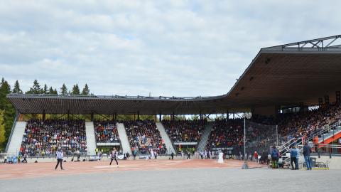 Pesäpallokentällä peli käynnissä etualalla ja takana näkyy katsomo, jossa väkeä istumassa.