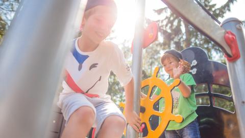 Kaksi lasta leikkii ulkona laivaa muistuttavalla leikkitelineellä. Vihreäpaitainen poika ohjaa keltaista ruoria, valkopaitainen poika arpoo, laskeutuuko metallista tankoa pitkin alas maahan.