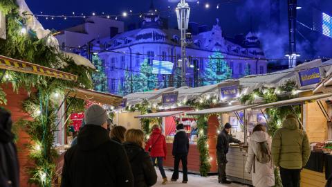 People at dusk in the Christmas market with Christmas decorations and illuminated wooden huts, with the building lit up with light art in the background.