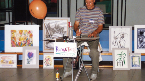 Henkilö istuu pöydän ääressä piirtämässä. Hänen ympärillään on erilaisia tauluja.