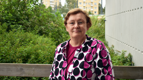 Henkilö seisoo rakennuksen edessä. Taustalla on pensas.