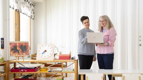 Two women looking at a laptop. On their left a shelf with miscellanous items, including children's toy clock.