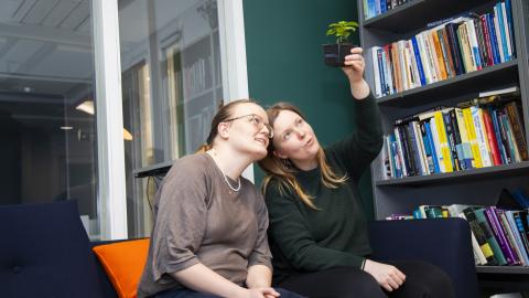 Two women sitting on the couch. The other one holds a small plant in a pot, which both are looking at. On the right there's a book shelf.