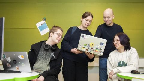 Neljä ihmistä, joista laitimmaiset istuvat pöytien ääressä. Yksi pitelee kannettavaa tietokonetta, jota kaikki katsovat.