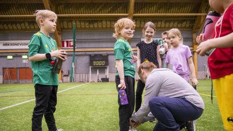 Lapsia rivissä urheiluvaatteissa nurmikentällä. Aikuinen sitoo yhden lapsen kengännauhoja.