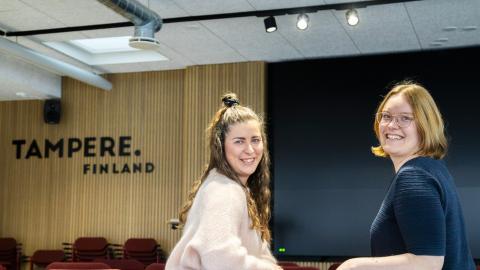 Kaksi henkilöä istuu punaisilla tuoleilla. He ovat kääntäneet katseensa kuvaajaan päin. Taustalla on teksti Tampere.Finland.