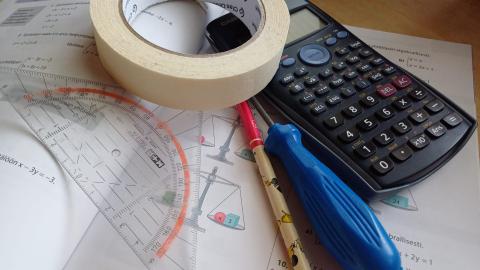 Matematiikan kirjan päällä laskin, maalarinteippiä, ruuvimeisseli, kynä ja piirtokolmio.