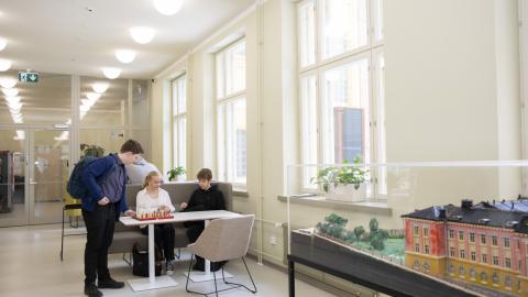 Kolme nuorta juttelee keskenään pöydän ääressä. Kuvassa oikealla on koulurakennuksen pienoismalli. 