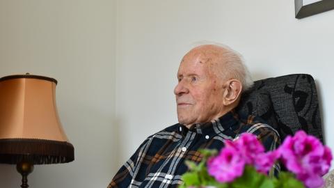 Henkilö istuu nojatuolissa. Hänen edessään on kukkakimppu ja vieressä pöytälamppu.