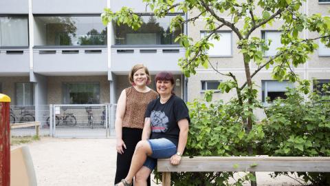 Kaksi työntekijää on kerrostalossa sijaitsevan päiväkodin pihalla. Edessä on leikkivälineitä ja takana kerrostalo.