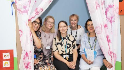 Viisi naista istumassa valkoisten, vaaleanpunakuvioisten verhojen välissä.