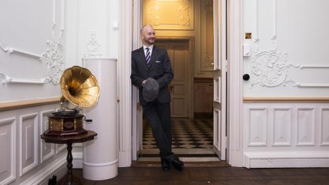 Henkilö seisoo hymyillen nojaten oven pieleen. Oven vasemmalla puolella katsojasta nähden on pienellä pöydällä gramofoni.