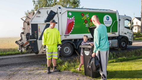 Jäteauto noutaa jätteet jäteastiasta. Etualalla kuljettaja ja mies ja poika, joilla mukana jätesäkki.