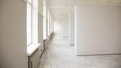 The large gallery space has windows on one wall and three white walls.
