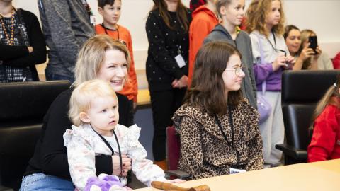 Hymyilevä äiti istuu pöydän ääressä pikkulapsi sylissään. Pöydän ääressä ja takana on myös muita lapsia.