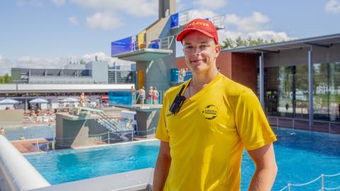 A life guard is standing at an outdoor pool with a diving platform behind him.