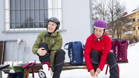 Kaksi kypäräpäistä henkilöä istuu ulkona penkillä ja vaihtaa luistimia.