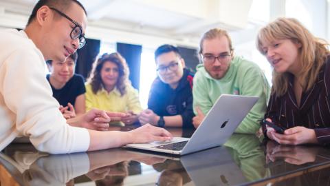 Kuusi henkilöä iloisesti katsomassa tietokoneen näyttöä valoisassa toimistotilassa.