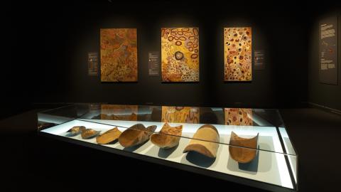 Hämärässä näyttelytilassa on seinillä värikkäitä maalauksia. Edessä lasivitriinissä puisia esineitä.