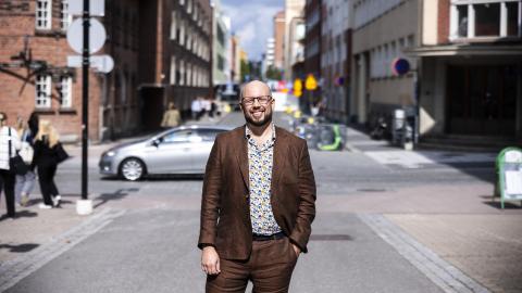 Mies seisoo kadulla hymyillen ja katsoo kameraan. 