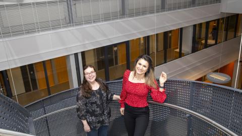 Kaksi henkilöä hymyilee iloisina metallisissa portaissa. Toinen vilkuttaa.