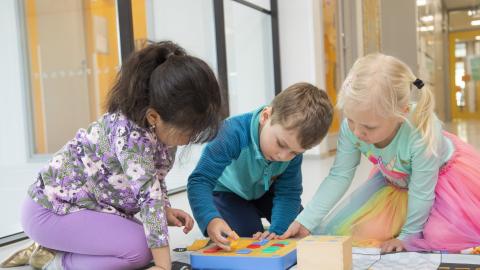 Three kindergarten-age children are sitting on the floor in front of a large window, playing with blocks.