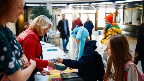A city employee offers sweets to a child over the presentation table. The child is wearing a funny light blue animal hat. Other children and adults can be seen in the background.