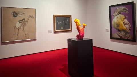 Näyttelytila, jossa on valkoiset seinät ja punainen matto. Seinillä kolme maalausta ja edessä oranssi kukkamainen veistos. 