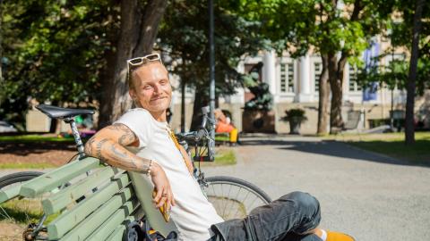 Man sitting casually on a park bench, smiling, big trees in the background.