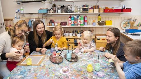 Three adults and four small children at a big table. The children are looking intently at the colourful paint cans. In the background, a shelf of neatly arranged art supplies can be seen.