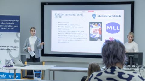 Kaksi henkilöä seisoo luentotilan etuosassa ja taustalle on heijastettu kuva tilaisuuden aiheesta.