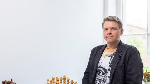 Mies istuu shakkilaudan äärellä, katsoo kameraan.