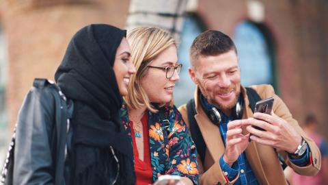 Kolme hymyilevää ihmistä istuu rinnakkain. Yksi heistä näyttää muille jotakin puhelimen näytöltä.