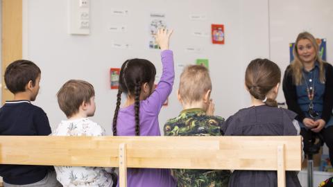 Lapsia istuu penkillä ja yksi heistä viittaa. Aikuinen istuu edessä.