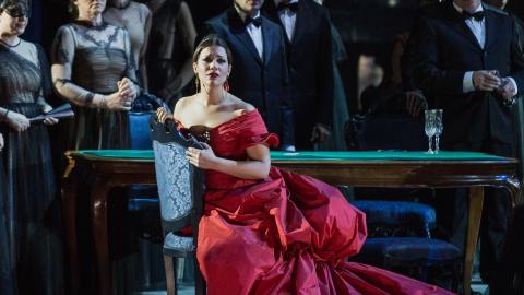 Nainen punaisessa iltapuvussa istuu tuolilla ja katsoo eteenpäin. Taustalla ihmisiä juhla-asuissa.