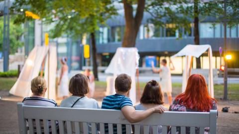 Viisi henkilöä istuu Hämeenpuistossa penkillä ja katsoo edessä olevaa taide-esitystä.
