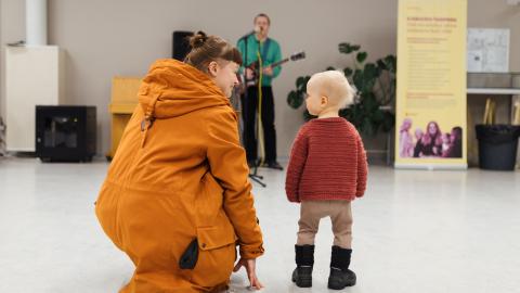 An adult is crouched next to a small child in the Tampere Residents' Evening. They look at each other. In the background, a musician stands out, playing a guitar.