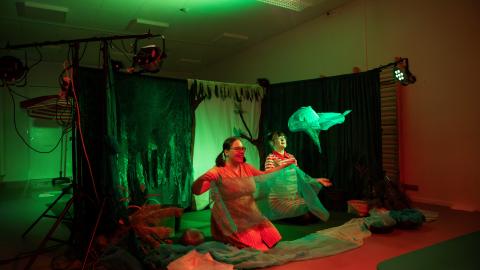 Käynnissä on Metsäretki-niminen esitys, jossa vihreät lavasteet ja kaksi esiintyjää heiluttelee sinisiä huiveja. 