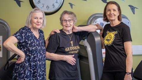Kolme seniori-ikäistä hymyilevää rouvaa sseisoo vierekkäin kuntosalilla.