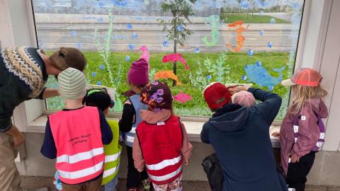 Kuusi värikkäisiin huomioliiveihin pukeutunutta lasta piirtää ikkunaan ikkunatusseilla yhdessä kahden aikuisen kanssa.