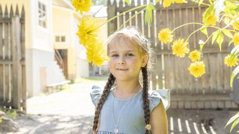 Pikkutyttö hymyilee kameralle, keltaisten kukkien joukosta. Taustalla museokorttelin taloja ja aitaa.