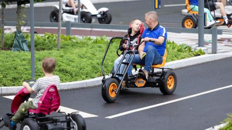 In a traffic park, children pedal on a pedal cars, in the middle  a parallel pedal car with a child and an adult.